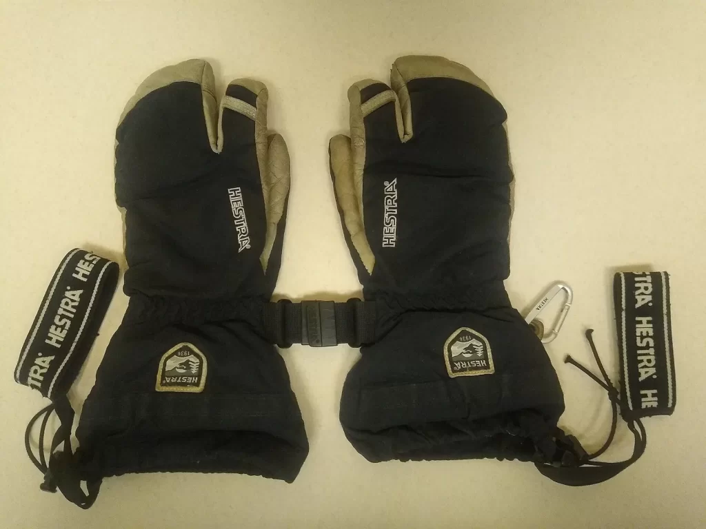 Une paire de gants de ski Hestra à trois doigts, bleu marine à paume en cuir, posée à plat avec ses dragonnes