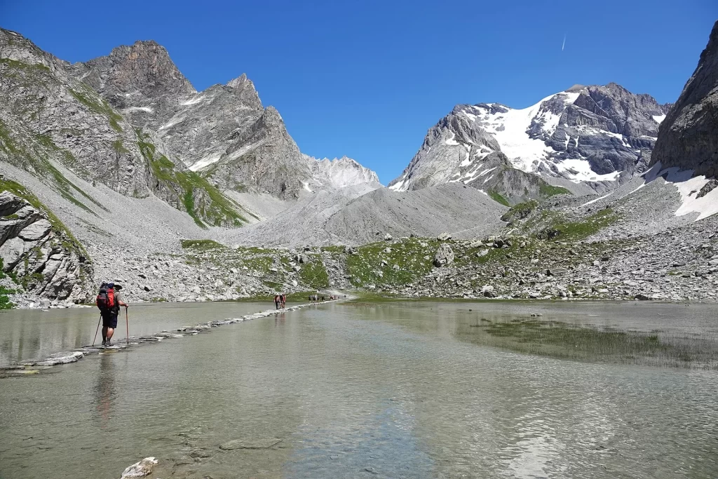 Randonneur vu de dos, sac à dos et bâtons, traversant le lac des Vaches sur un chemin de dalles face à la Grande Casse